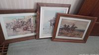 Three Western-themed framed art prints leaning against a wall showing scenes with horses and riders, each with wooden frames and white matting.