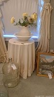 Round table covered in white ruffled cloth, ceramic pitcher and basin with artificial yellow roses placed on it, large glass jug in front, framed landscape painting propped against wall.