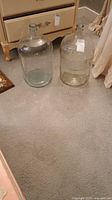 Two large clear glass jugs side by side on a carpeted floor near furniture.