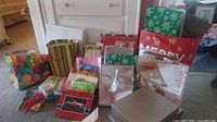Wide shot showing an assortment of colorful holiday gift bags with different designs like stripes, snowflakes, and holiday messages alongside multiple gift boxes in neutral colors.