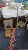 Photo showing a cream metal plant stand with three circular wire mesh shelves and decorative scrollwork. Next to it are three framed wall hangings with artwork of a cat and two butterflies laid on the floor.