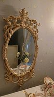 Gold-tone ornate oval mirror with decorative swirl frame hanging on wall.