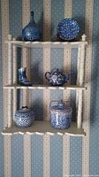 Full view of shelf containing six blue and white ceramic items including vase, boot, plate, teapot, and two covered jars.