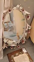 Oval mirror with white metal leaves and pink flowers on the frame, hanging near beige curtains and a beige chair visible in the reflection.