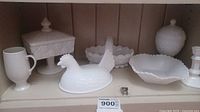 Full assortment of six white milk glass pieces arranged on shelf including chicken candy dish (bottom missing), lidded pedestal dish, footed mug, scalloped bowl, lidded urn shaped piece.