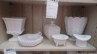 Full shelf view showing six assorted milk glass items including vases, bowls, covered jar, and footed basket with various decorative patterns.