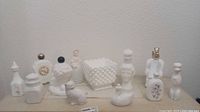 View of 10 white milk glass Avon perfume bottles of various shapes and a square hobnail milk glass candy dish with scalloped edges.