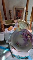 Photo showing a collection of items including floral framed art, brass candlesticks, vintage plate with floral design, carved bird figurines, hand-stitched linens, and ceramic dish.