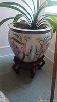 Front view of round ceramic flower pot with colorful Asian floral designs and glossy finish, sitting on a dark carved wooden stand inside a room.
