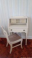 Front view of white painted secretary desk with multiple storage cubbies, single drawer, and matching wooden chair with striped seat in orange carpeted room.