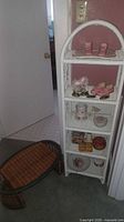 White rattan shelf unit with five shelves holding various small decorative items and a brown woven tray beside it.