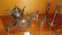 Overall view of silver plated tea pot, candle holders, butter dish lid, round plate, and small perforated candle holder on wooden shelf.