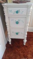 White wooden cabinet with three drawers painted with green leaves and turquoise knobs sitting on orange carpet.