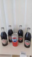 Four vintage Coca-Cola glass bottles and one vintage Pepsi can displayed on a white surface, showing front views of labels.