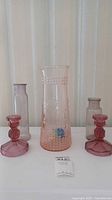 Full view of the lot showing two cranberry glass candle holders, one tall pink glass pitcher, and two small clear bottles on a white surface.