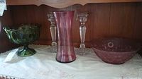 Photo showing green glass candy dish on pedestal, cranberry vase, cranberry bowl with floral pattern, and two crystal candle holders on white embroidered cloth.