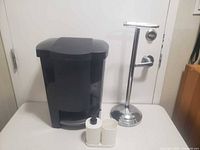 Black plastic trash can, chrome toilet paper stand, white soap dispenser, tumbler and tray on tabletop