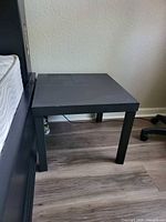 Black side table positioned next to a bed and near a chair on a wooden floor.