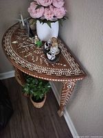 Photo showing the half-moon wooden accent table with intricate white inlay patterns on the top surface and legs, decorated with floral and geometric designs. Table positioned against wall with decorative items displayed on top.