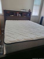 Full queen size bed frame in dark brown wood with built-in headboard featuring open storage shelves. Mattress included, white quilted surface visible.