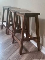 Set of three wooden bar stools arranged side by side on a wood floor against a wall, showing the stools' classic dark wood finish and simple design.