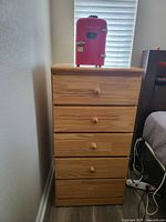 Photo of a solid wood dresser with five drawers and round wooden knobs, light-toned wood, standing between a wall and bed with a window behind it.
