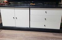 Photo showing front view of TV stand featuring three white drawers on right and double-door cabinet on left, black knobs, dark top surface, placed on wood floor.