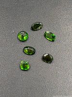 Six faceted chrome diopside gemstones displayed on dark surface showing their deep green color and faceting.