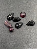 Seven garnet gemstones displayed on a gray background showing various shapes and colors including teardrop, round beads, and a rough crystal.