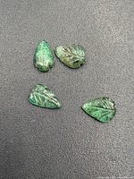 Four carved leaf-shaped jade gemstones featuring detailed vein carvings and varying green shades, arranged on a dark surface.