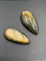 Two polished labradorite teardrop gemstones showing golden and green labradorescence on dark surface.