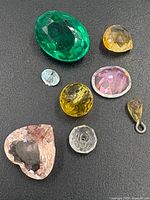 Nine mixed loose faceted gemstones on black background showing varied shapes and colors including green, pink, yellow, blue and clear stones.