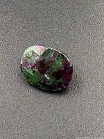 Faceted oval rubellite gemstone showing mixed green and reddish-purple colors on a textured dark background.