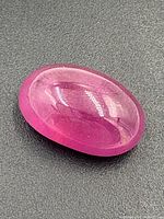 Oval cabochon ruby gemstone showing smooth polished surface with a bright pink to purplish-red color on gray background