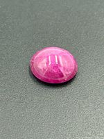 Close-up photo of a cabochon-cut ruby gemstone. The gem is a smooth, polished oval with a deep pink to reddish color and internal natural characteristics.