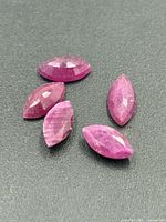 Photo showing five marquise-shaped faceted ruby gemstones in shades of pink on a dark surface, highlighting their shape and color.