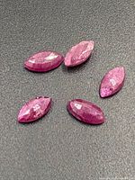 Five marquise cut ruby gemstones arranged on a dark surface showing pinkish-red color and faceted cuts.