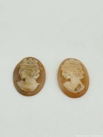 Photo showing two oval shell cameos with female profile busts carved in relief, on plain background.