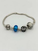 Full view of the sterling silver snake chain charm bracelet with three distinct beads: a silver patterned bead, a blue glass bead, and a silver charm with a cross and blue accents.