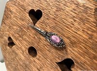 Close-up angled view of the vintage pin showing the pink cat eye glass cabochon set in ornate silver-tone metal casing, placed on a wooden surface with heart-shaped cutouts.