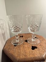 Two clear crystal glasses placed on a wooden surface with heart cut-outs, showing front view of the cut pattern and faceted stems, with light reflections.