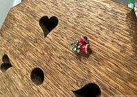 Close-up photo of a Christmas pin resting on a wood surface with heart-shaped cutouts, showing front side of the pin with red bells and green holly.