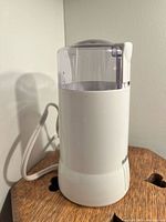 Front view of white Salton electric coffee grinder with clear plastic lid and attached power cord, on wooden table against white wall.