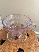 Clear plastic coffee dripper with ribbed, conical shape and handle, purple tinted, placed on wooden surface