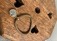 Gold-coloured bracelet displayed on wooden surface with visible heart-shaped vintage charm and round tag charm.