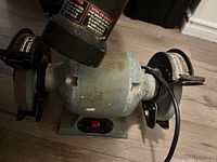 Bench grinder showing the body, power switch, and two buffing wheels with protective guards, visible rust on metal parts.