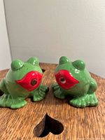 Two green ceramic kissing frog salt and pepper shakers with red lips on wooden surface.