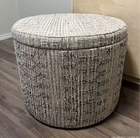 Photo of a large round storage ottoman or toy box with textured woven fabric upholstery in neutral colors.
