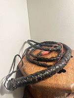 Close-up photo of black braided leather bull whip coiled on wooden surface against white wall. The handle has a loop and the whip shows a small red fiber detail near the handle.