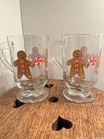 Four clear glass mugs with gingerbread men and candy cane Christmas decoration, two of one style and two of another style, showing differing bases and handles.